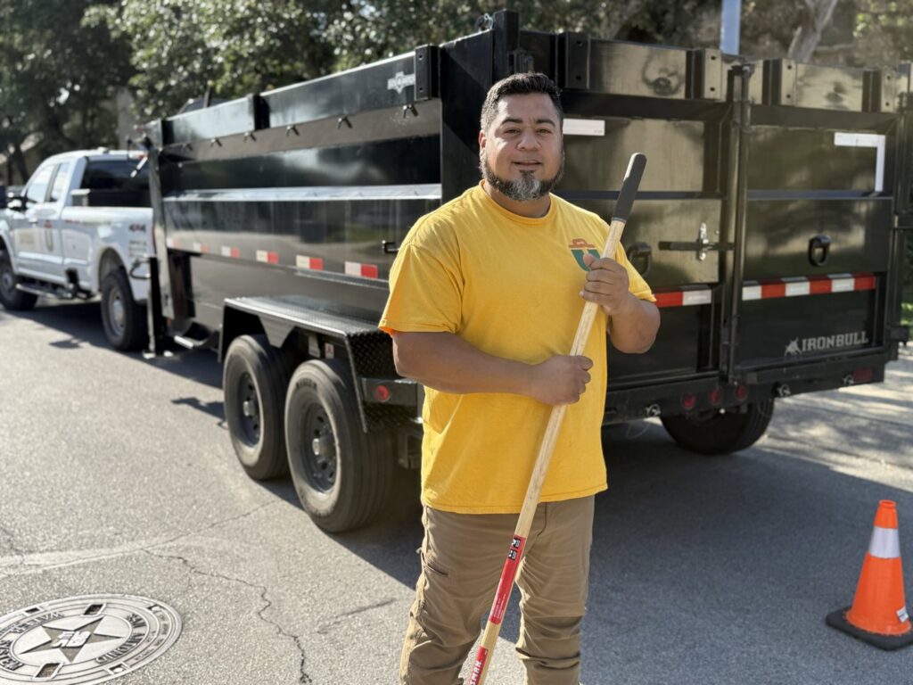 A Silverhull Junk Removal worker in a yellow shirt holding a tool, standing in front of their dump trailer in San Antonio, TX