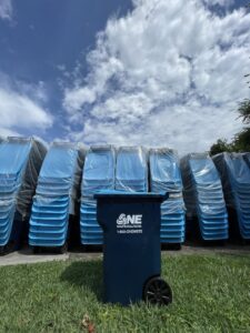 A single blue trash bin from One Waste Solutions in the foreground with a stack of new bins behind it in Murfreesboro, TN.
