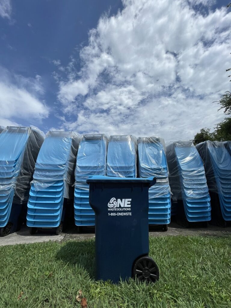 A single blue trash bin from One Waste Solutions in the foreground with a stack of new bins behind it in Murfreesboro, TN.