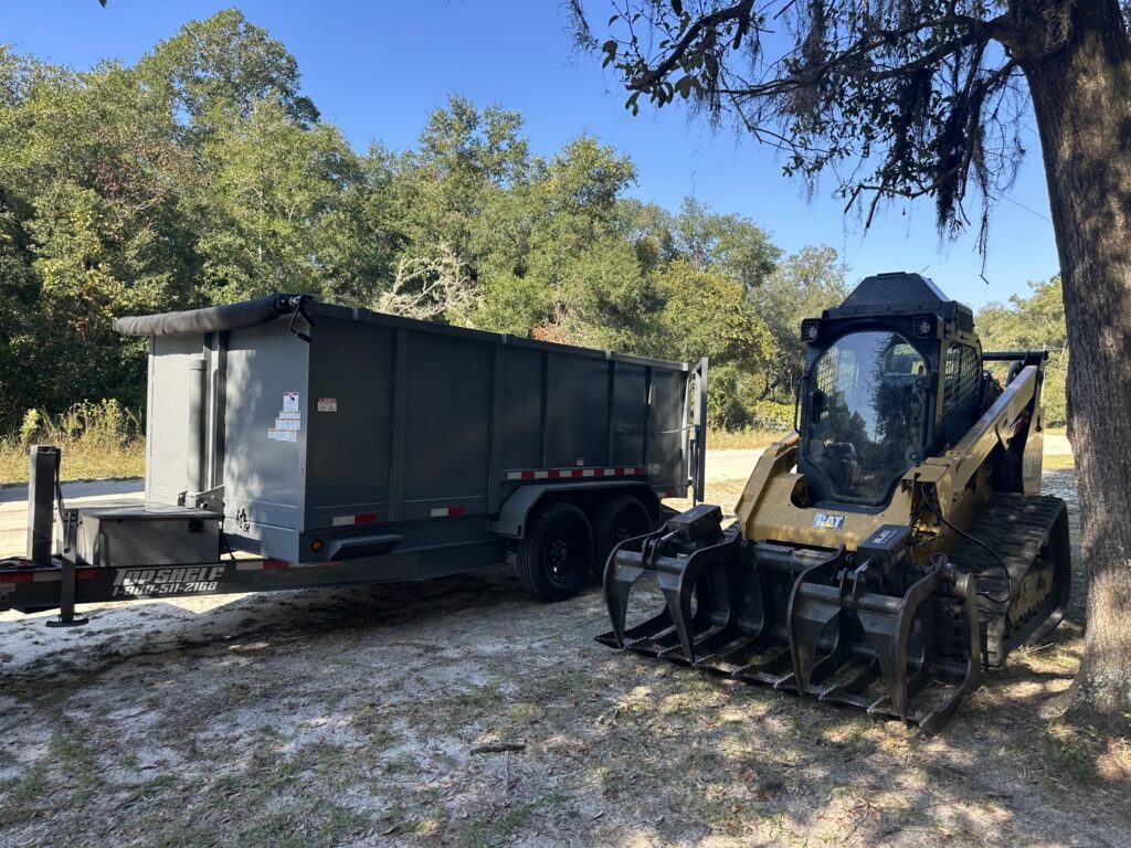 A skid steer parked next to a gray dumpster trailer from Big Skye Dumpsters, ready for junk removal in Jacksonville, FL.