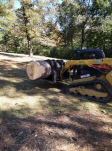A skid steer loader with a grapple attachment efficiently hauling a large tree log, demonstrating services by Log and Leaf Tree Service in Hoover, AL.