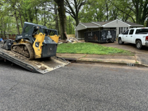 A skid steer loader on a trailer at a Dts dumpster rentals junk removal site in Memphis, TN, with a full dumpster.