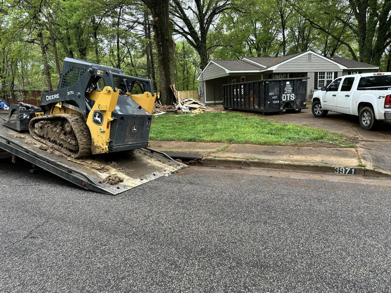 A skid steer loader on a trailer at a Dts dumpster rentals junk removal site in Memphis, TN, with a full dumpster.