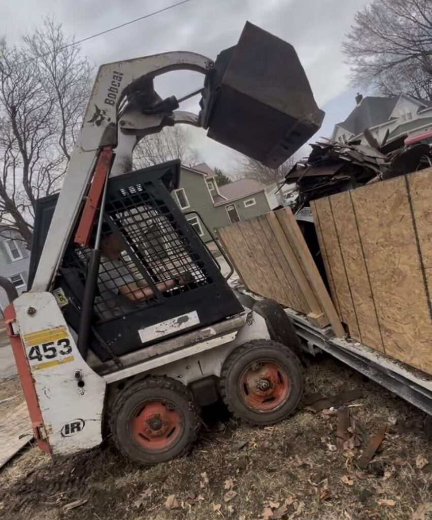 A skid steer loading construction debris into a large container for Rosales Junk Removal in Des Moines, IA.