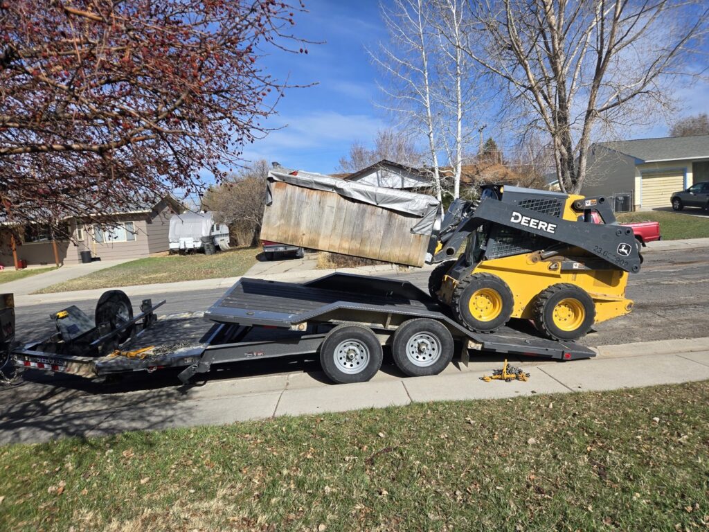 A yellow skid steer loading a large wooden container onto a flatbed trailer for Rubble Removers LLC in Riverton, WY.