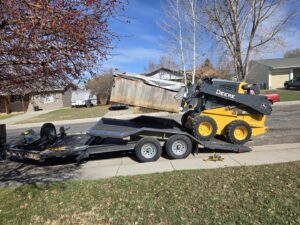 A yellow skid steer loading a large wooden container onto a flatbed trailer for Rubble Removers LLC in Riverton, WY.