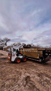 A skid steer loading a pile of debris into a dump trailer for efficient junk removal by Bin Suave LLC in Espanola, NM.