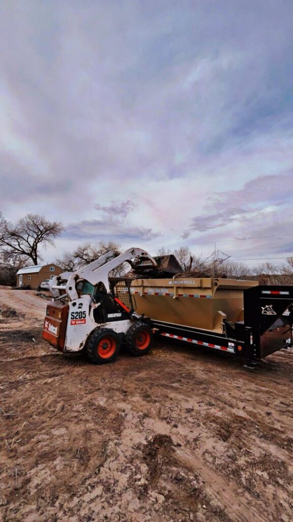 A skid steer loading a pile of debris into a dump trailer for efficient junk removal by Bin Suave LLC in Espanola, NM.