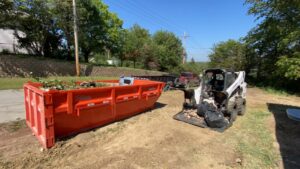A skid steer loader moving debris into a large orange roll-off dumpster for LNK Junk Removal in Council Bluffs, IA.