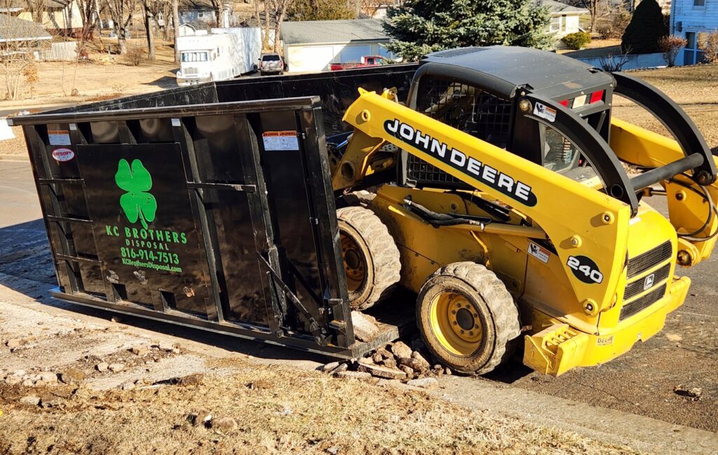 A John Deere skid steer loading debris into a KC Brothers Disposal dumpster during a junk removal job in Kansas City, MO.