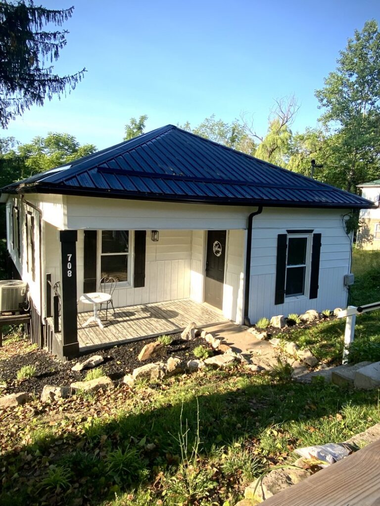 A small white house with a new black metal roof and updated landscaping, showcasing exterior renovation by In Goode Hands LLC in Beckley, WV.