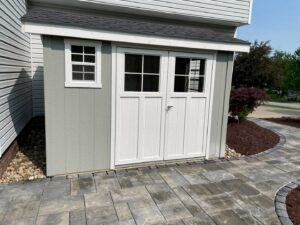 A newly installed small shed with white doors and a window, adjacent to a paved walkway by JDM Outdoors of Robinson in Pittsburgh, PA.