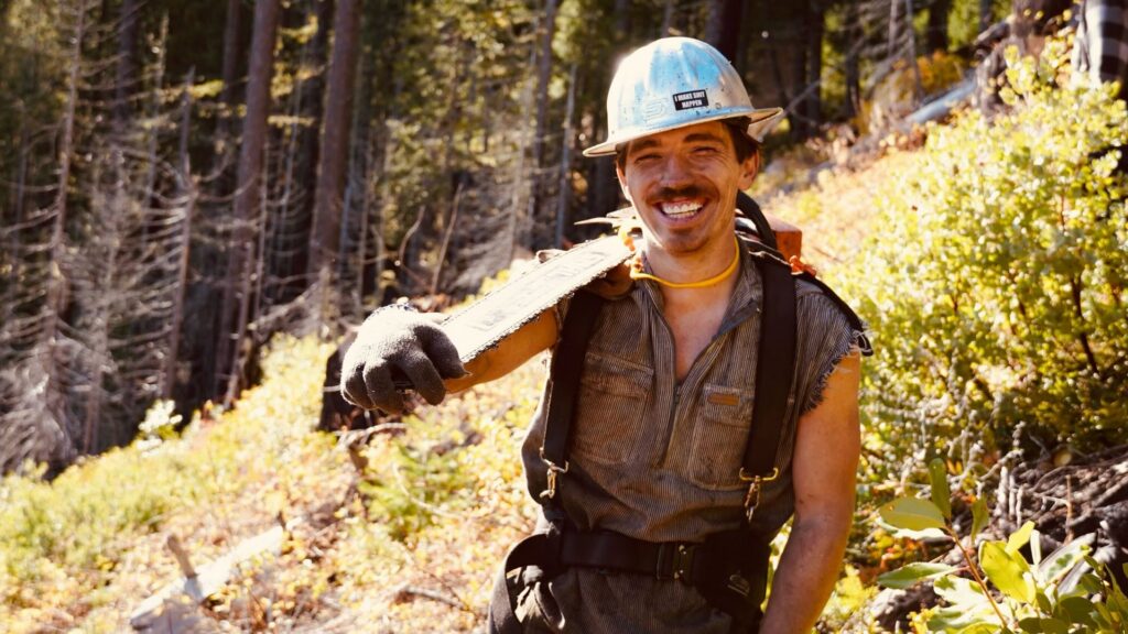 A smiling arborist from Wild Roots Arborist holding a chainsaw on his shoulder in a forest setting in Fayetteville, AR.