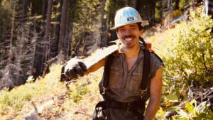 A smiling arborist from Wild Roots Arborist holding a chainsaw on his shoulder in a forest setting in Fayetteville, AR.