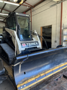 A compact track loader with a snow plow attachment on a trailer, representing snow removal services by Water Wizards Irrigation, Inc. in Enid, OK.