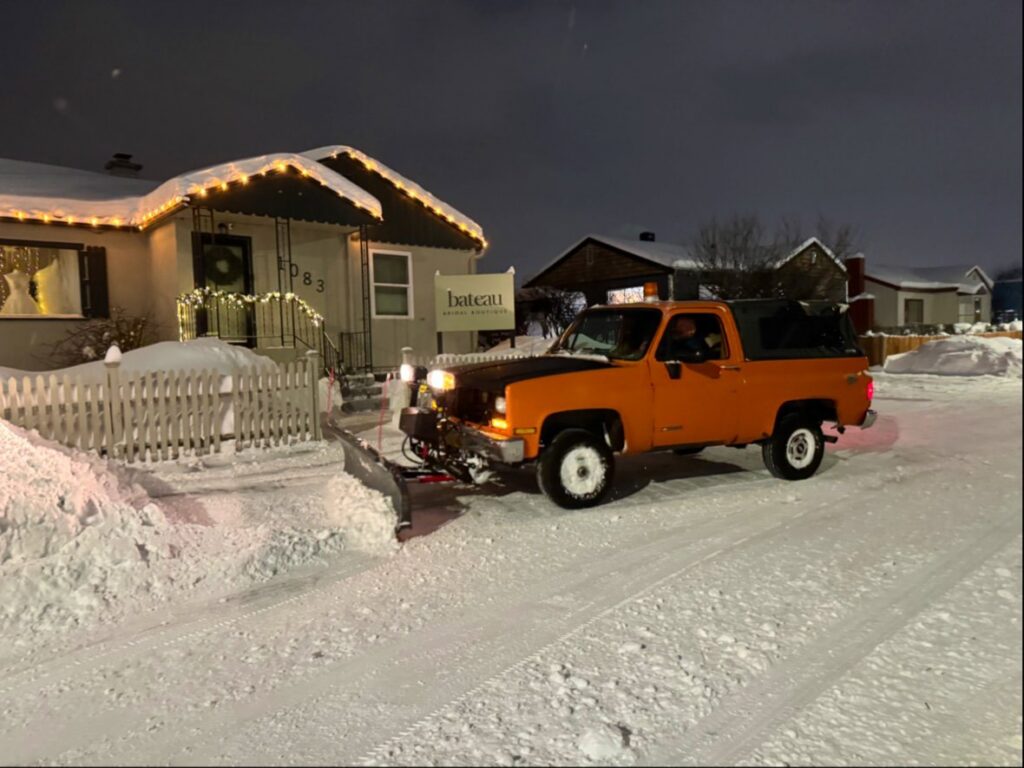 An orange snowplow truck from AK Property Maintenance, Inc. is clearing snow from a street at night in Anchorage, AK.