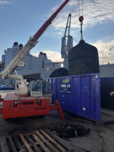 A crane loading a large, specialized item into a Toss-It dumpster near a dock or marina in Glen Burnie, MD.