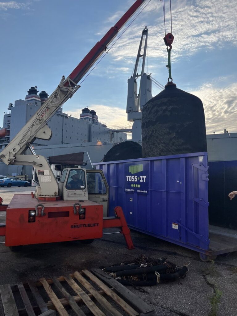 A crane loading a large, specialized item into a Toss-It dumpster near a dock or marina in Glen Burnie, MD.