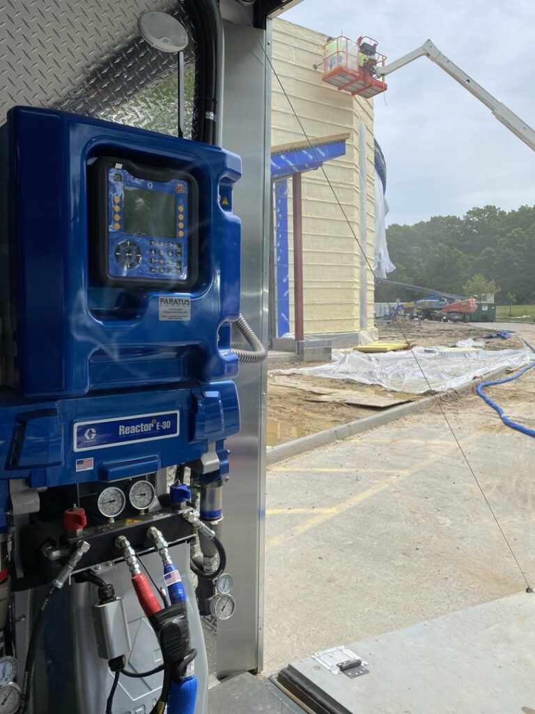 Spray foam application equipment inside a trailer, with a commercial construction site visible in the background by Olympus Spray Foam in Philadelphia, PA.