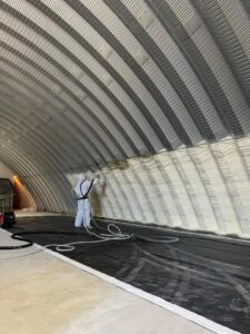 A worker applying spray foam insulation to the curved interior of a Quonset hut for Off-Axis Spray Foam and Radon Services in Clarksville, TN.
