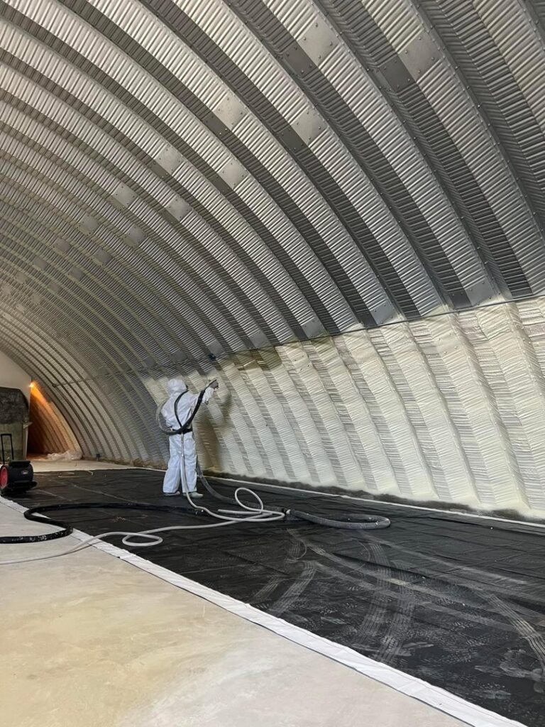 A worker applying spray foam insulation to the curved interior of a Quonset hut for Off-Axis Spray Foam and Radon Services in Clarksville, TN.