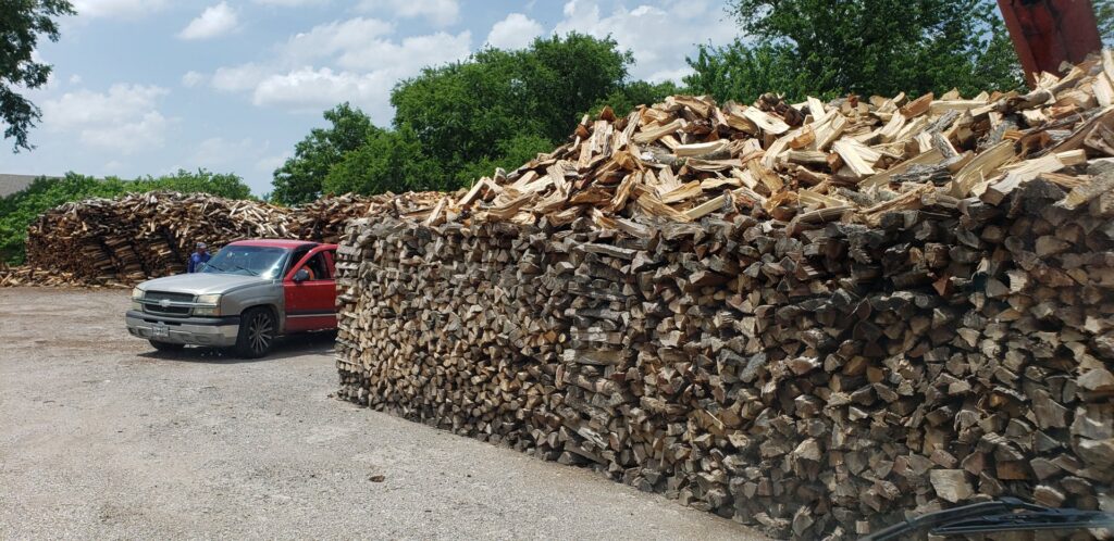 Large, neatly stacked piles of firewood and logs with a red pickup truck in the background at Personal Touch Tree Service in Dallas, TX