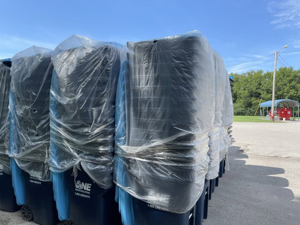 Stacked new blue trash bins, wrapped in plastic, with the One Waste Solutions logo in Murfreesboro, TN, ready for distribution.