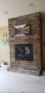 A beautifully installed stacked stone fireplace with a wooden mantel and artwork by The Foundation Guys in Lakewood, CO