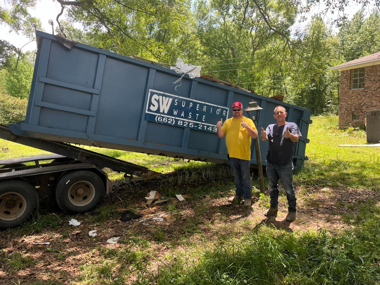 Two Superior Waste Management staff members giving thumbs up next to a blue dumpster being loaded for a junk removal job in Amory, MS.