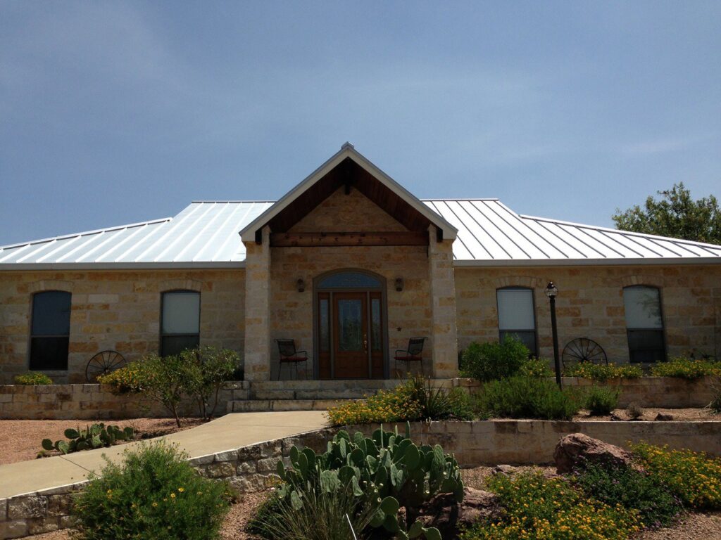 A beautiful single-story stone house featuring a new white metal roof and updated landscaping, completed by Custer Remodeling in Plano, TX.