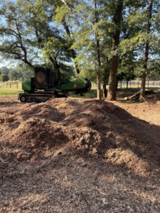 A green stump grinding machine visible behind a large pile of wood chips, demonstrating work by Manley's Stump Grinding in Opelika, AL.