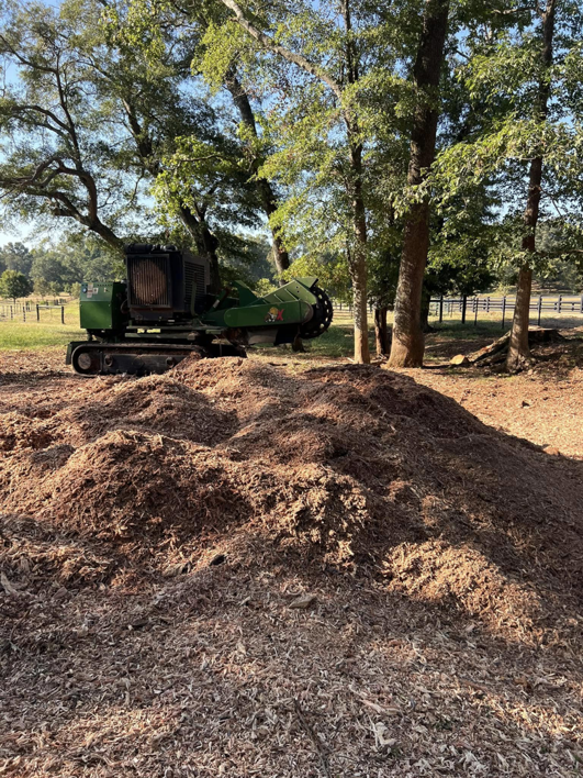 A green stump grinding machine visible behind a large pile of wood chips, demonstrating work by Manley's Stump Grinding in Opelika, AL.