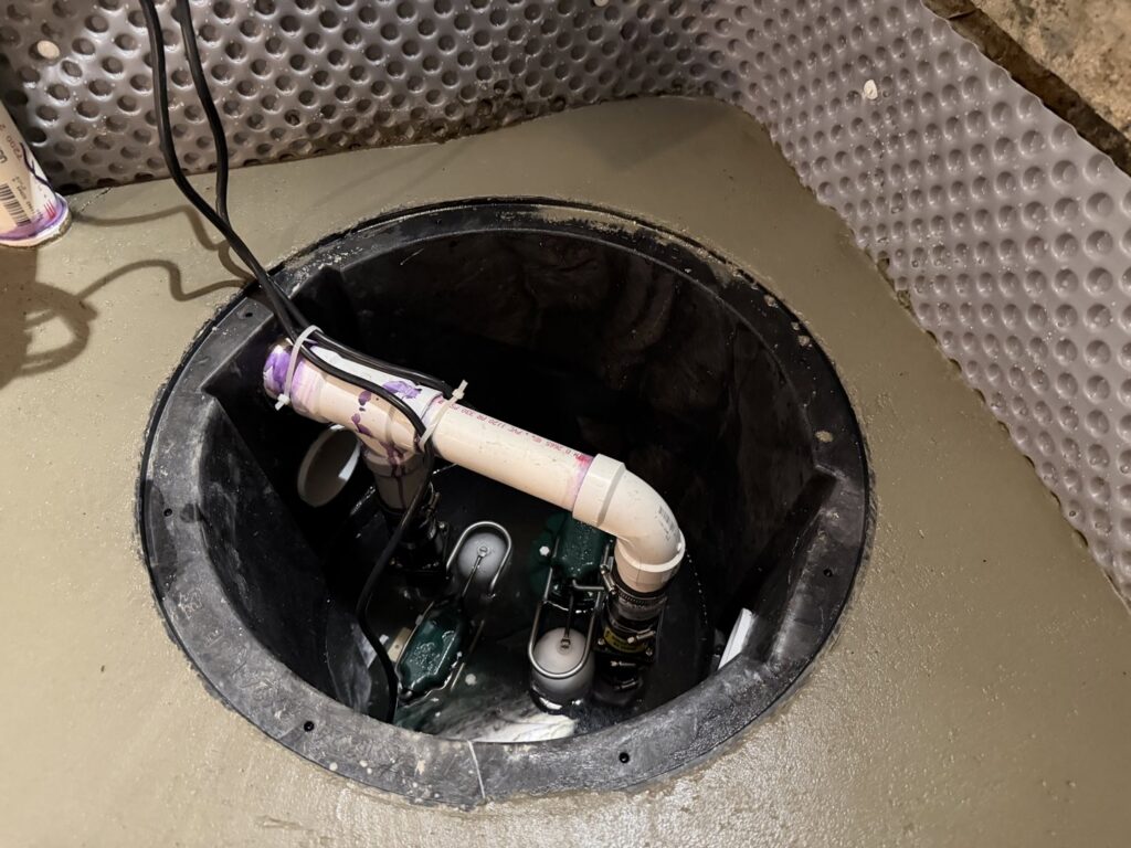 An installed sump pump pit with pipes and wiring, part of a basement waterproofing system by B-Dry System of New England in North Billerica, MA.
