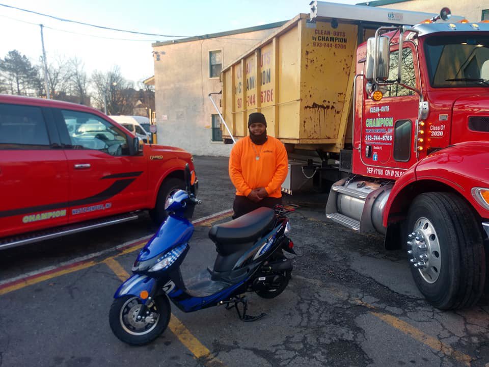 A Champion Waste Removal, Inc. team member standing with a blue scooter, with a junk removal truck and dumpster in the background in Orange, NJ.