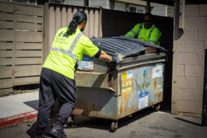 Two Bay Area Bin Support team members pushing a large commercial dumpster out of an enclosure in San Leandro, CA.