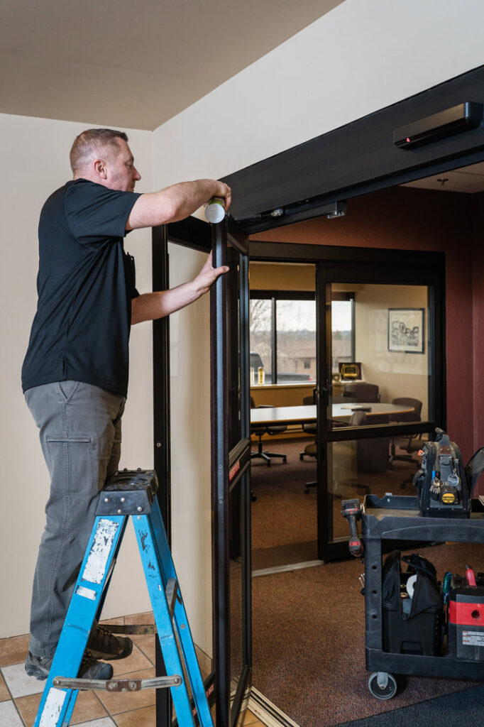 A technician on a ladder installing or repairing an automatic door for Automatic Entrances Of Wi in Waukesha, WI.