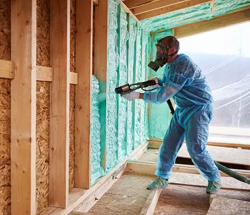 A technician in protective gear spraying green foam insulation onto a wall frame for Custom Insulation Company, Inc. in Worcester, MA