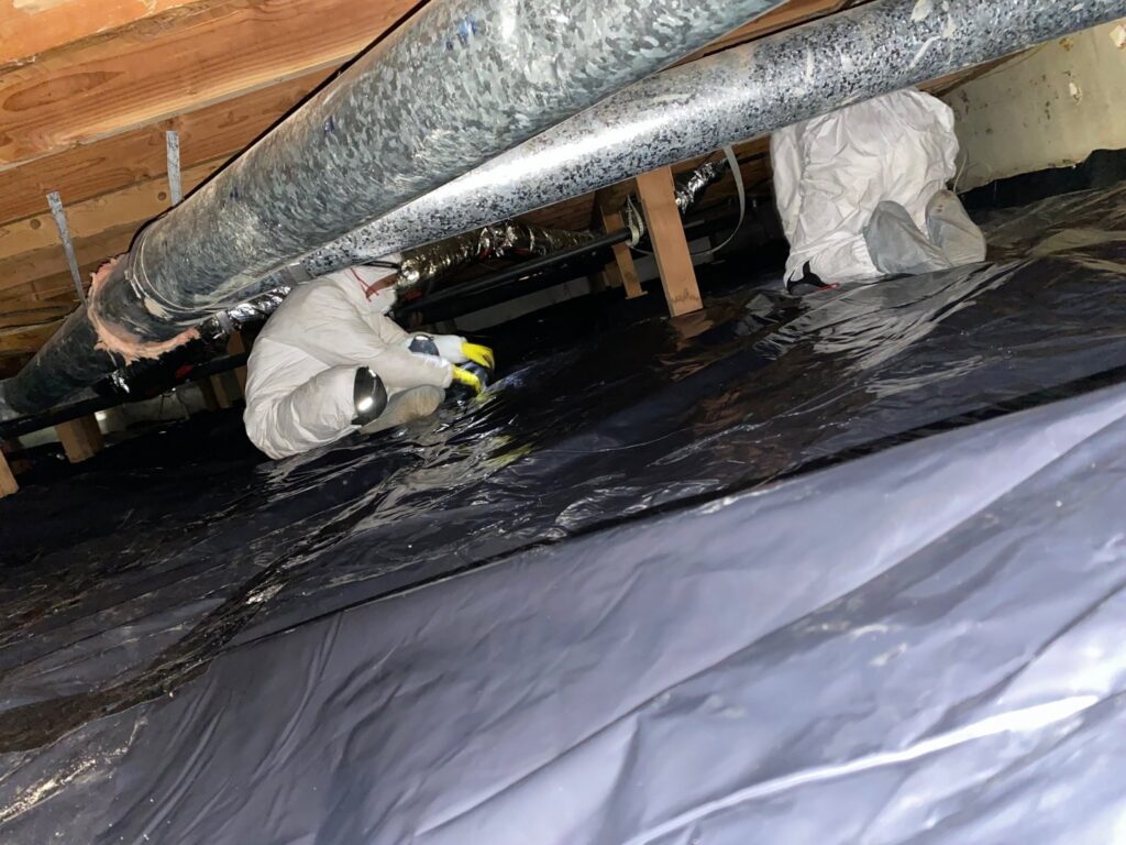 Two Northwest Crawl Space Services technicians in protective suits working on installing a black vapor barrier and large air ducts in a crawl space in Kent, WA.
