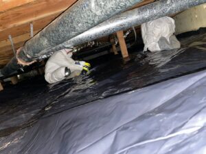 Two Northwest Crawl Space Services technicians in protective suits working on installing a black vapor barrier and large air ducts in a crawl space in Kent, WA.