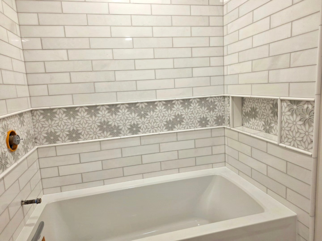 A tiled bathtub surround featuring white subway tiles and a floral accent strip by Tyler the Tiler in Jefferson, WI.