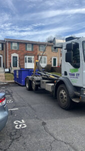 A Toss-It truck delivering a blue dumpster to a residential parking lot for a cleanout in Glen Burnie, MD.