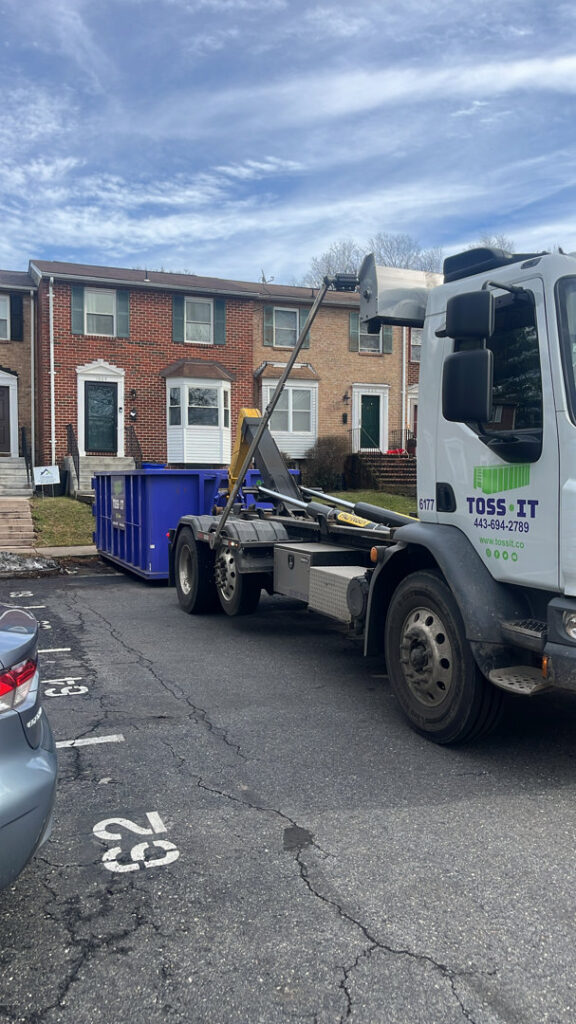 A Toss-It truck delivering a blue dumpster to a residential parking lot for a cleanout in Glen Burnie, MD.