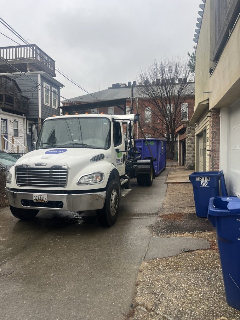 A Toss-It truck picking up a blue dumpster from a residential area after a junk removal job in Glen Burnie, MD.