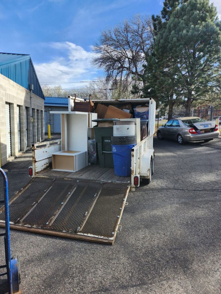 A trailer loaded with furniture pieces, bins, and a grill, parked near storage units for junk removal by Empty It Out Junk Removal, LLC in Billings, MT.