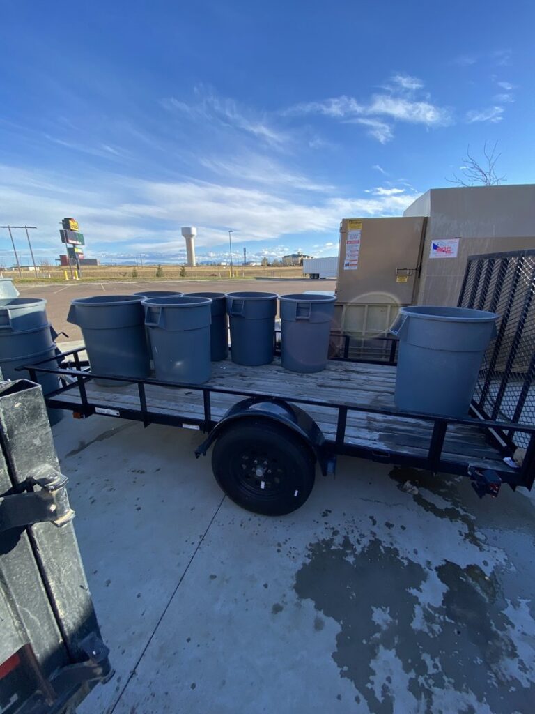 A utility trailer loaded with multiple gray trash cans, ready for transport by Hudson's Trash Removal, LLC in Great Falls, MT.