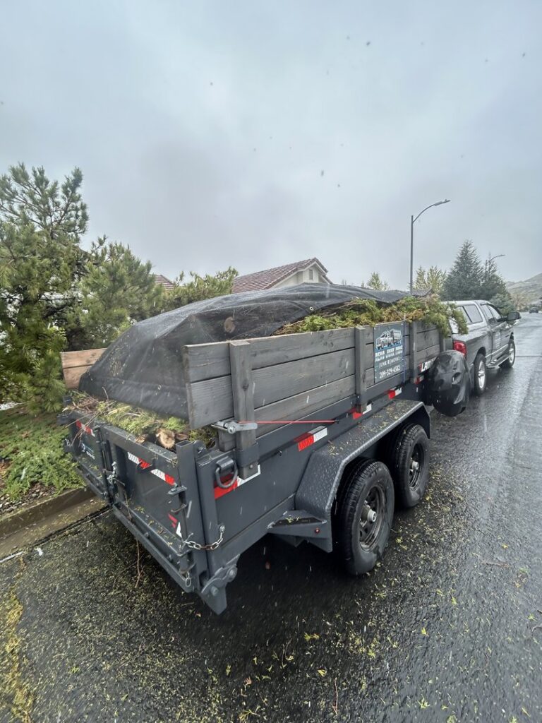A junk removal trailer loaded with green yard waste and covered with a tarp on a wet residential street by Reno Junk Pros in Reno, NV.