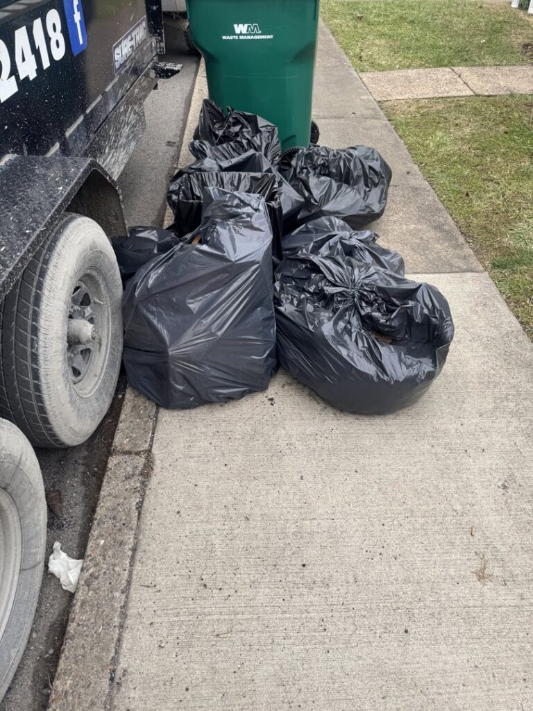 Black trash bags lined up on a sidewalk next to a Greybeard Dumpster Rentals trailer in Morgantown, WV, ready for pickup.