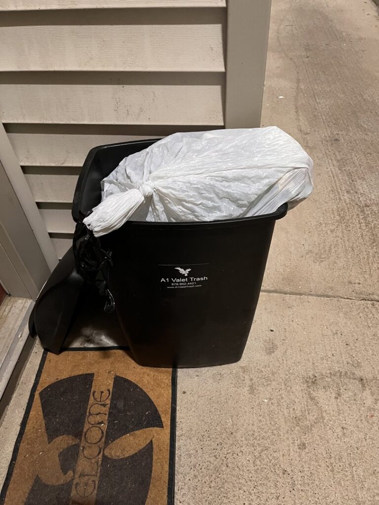 A black trash bin with a white trash bag inside, placed on a doorstep for A1 Valet Trash pickup in Atlanta, GA.