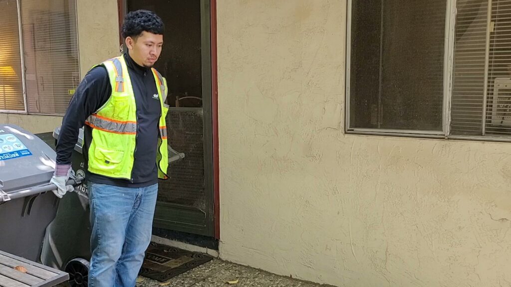 A TrashScouts employee in a safety vest and gloves moving a large waste bin in Oakland, CA.