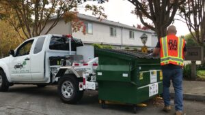 A TrashScouts employee in a safety vest servicing a green dumpster with a company truck nearby in Oakland, CA.